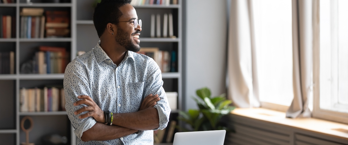 May 19 blog Young male business owner smiling while looking out office window.